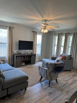 A living room with a ceiling fan, a flat-screen TV, a gray sofa, a patterned armchair, and a bookshelf with books and a Wi-Fi sign. - Hook, Relax & Stay: 3 BR Fishing Haven in Aransas Pass (Aransas Pass)