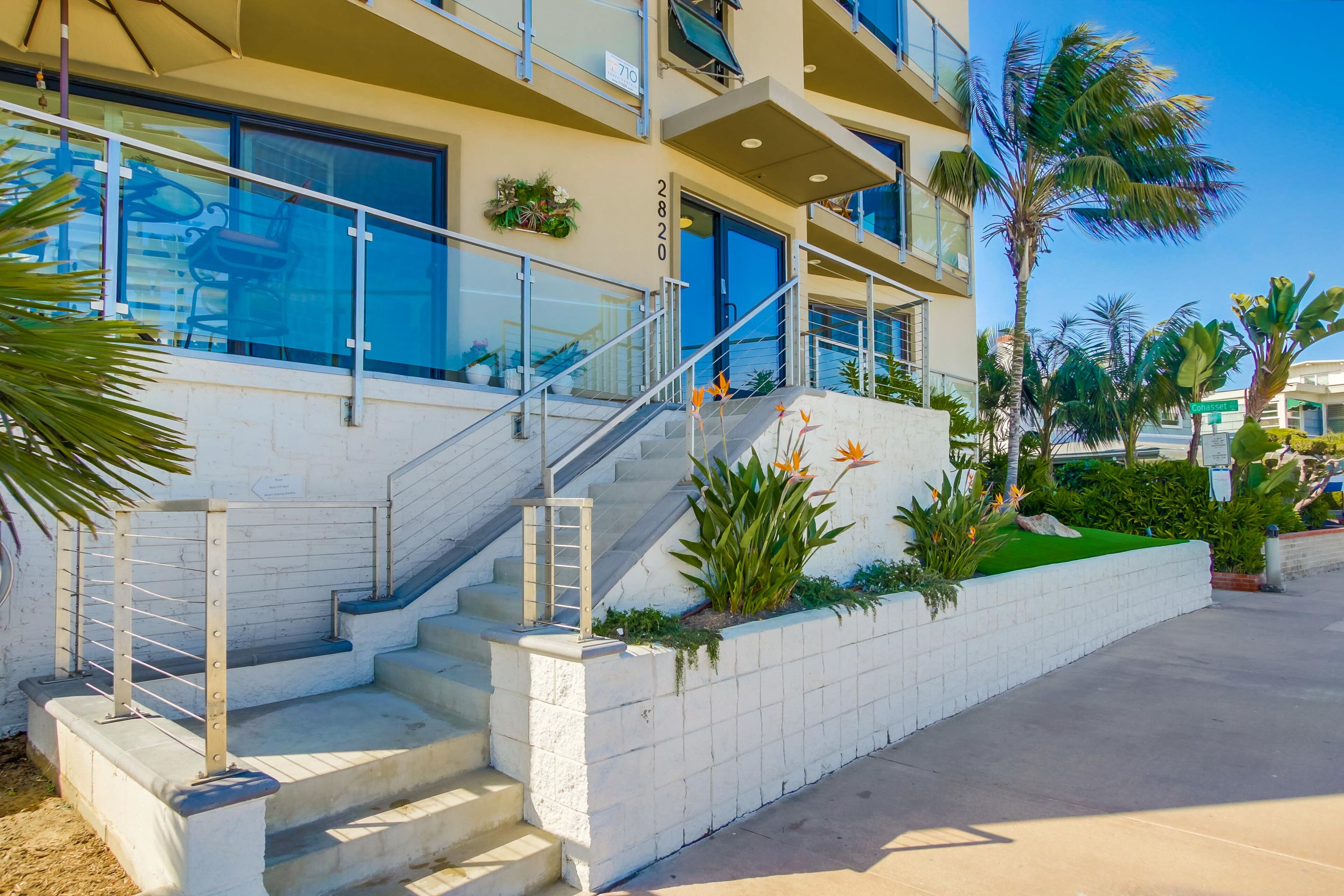 A multi-story building with a staircase leading to the entrance, surrounded by palm trees and a well-maintained garden.