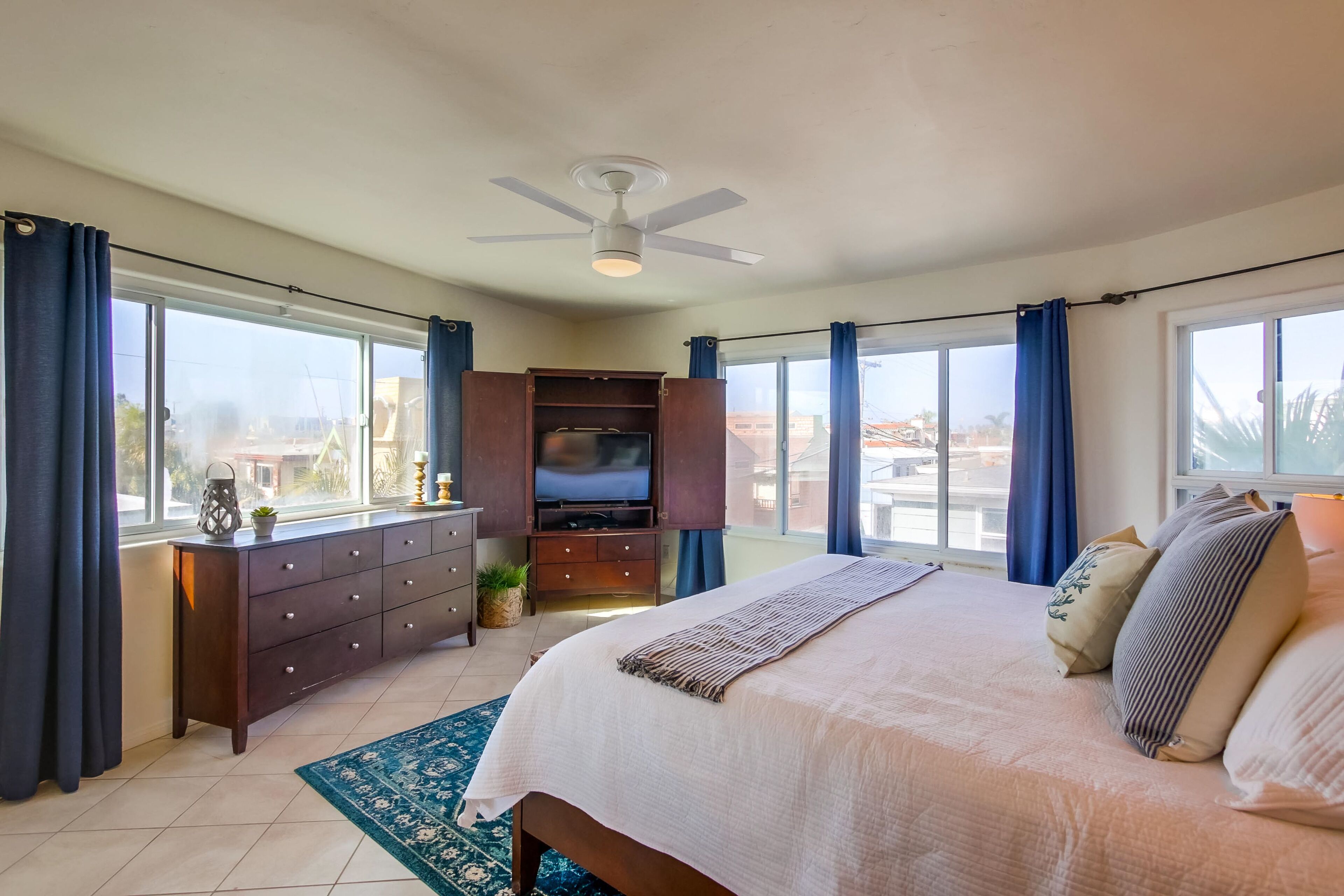 A bedroom with a large bed, a ceiling fan, a TV, and a view of the city through large windows.