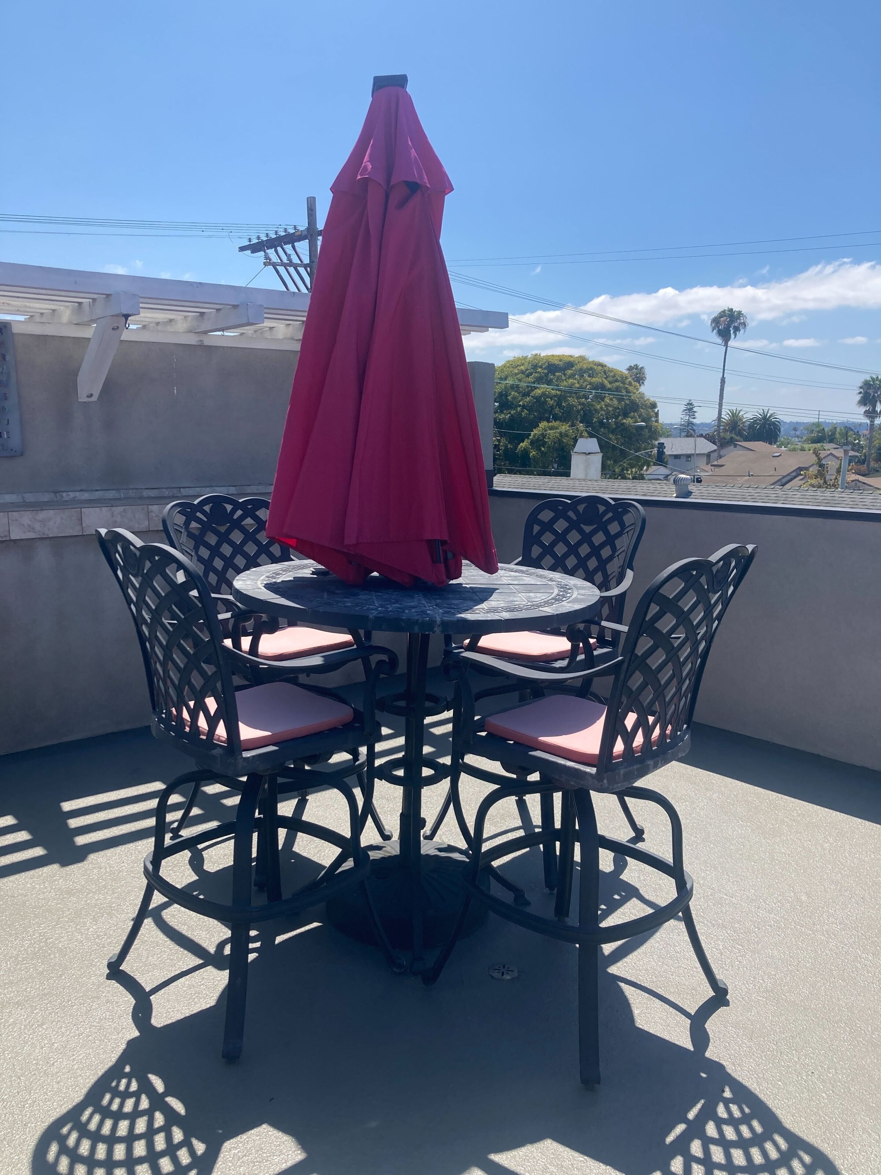A round table with a red umbrella, surrounded by black chairs with pink cushions, on a rooftop terrace with a view of the ocean.