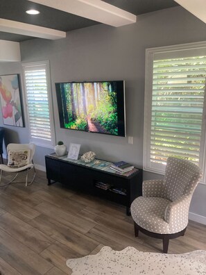 A living room with a flat-screen TV, a patterned armchair, a white chair, a console table with books, and a large window with blinds. - Ingraham Delight Pacific Beach (San Diego)