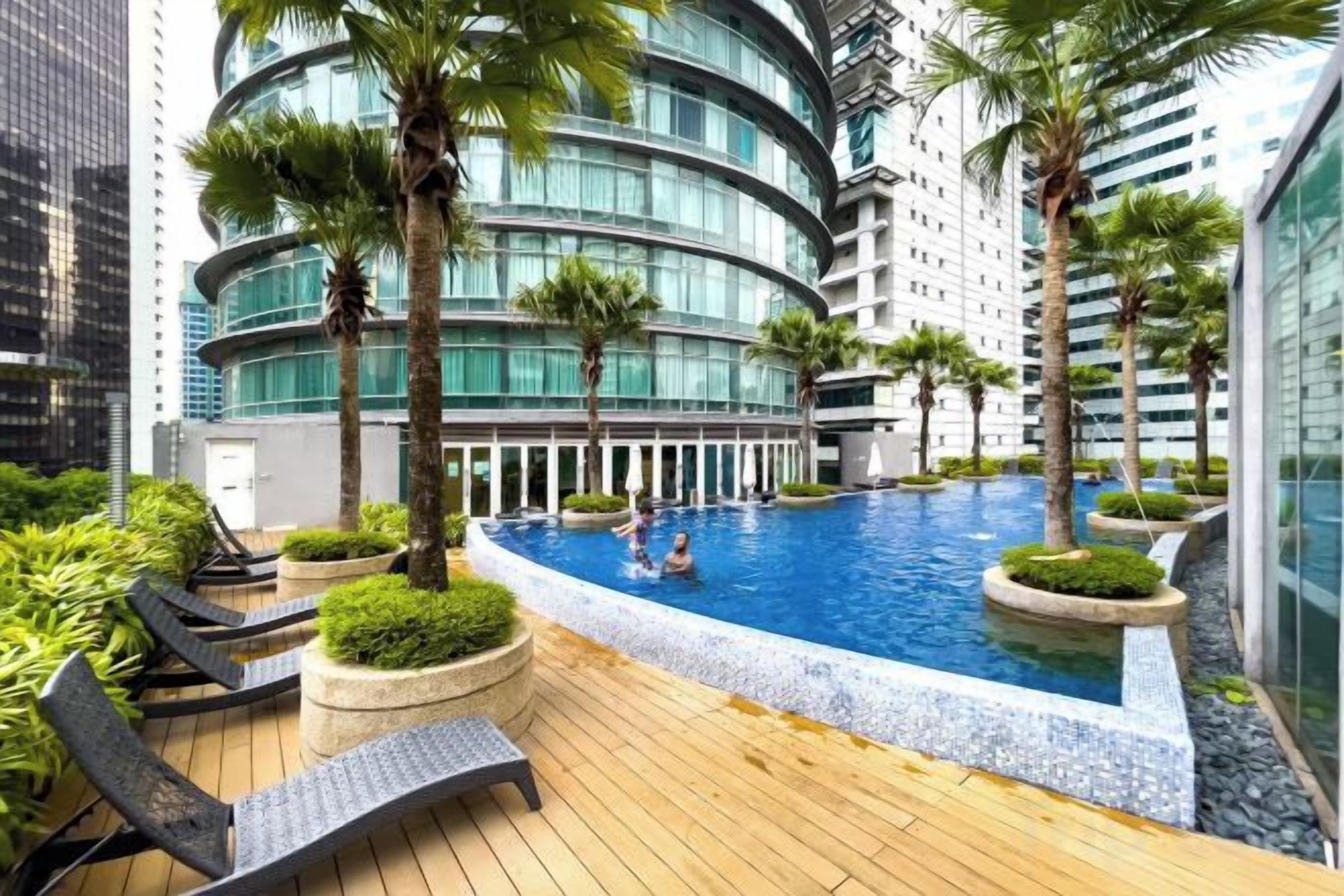 A swimming pool surrounded by lounge chairs and palm trees, with modern buildings in the background.