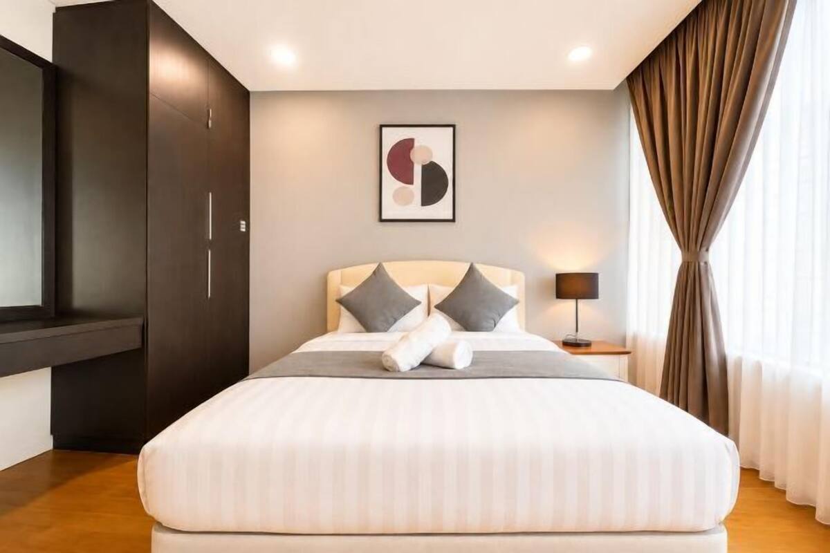 Un lit bien fait, avec des oreillers, une table de chevet avec une lampe, une armoire sombre et une œuvre d’art accrochée au mur.