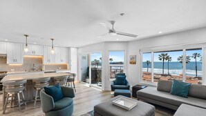 Eine moderne Küche mit weißen Schränken und einer großen Kochinsel, ein Essbereich mit Barhocker und ein Wohnzimmer mit Blick auf den Strand.