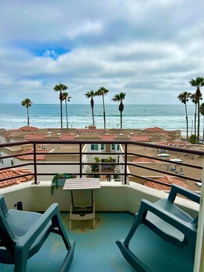 A balcony with two rocking chairs, a small table, and a view of the beach with palm trees and buildings.