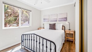 Une chambre à coucher comprenant un lit, une table de chevet, une lampe de chevet, un ventilateur de plafond et une fenêtre donnant sur des arbres.