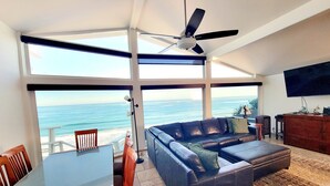A living room with a large glass door leading to a balcony with a view of the ocean. There is a ceiling fan, a TV mounted on the wall, and a leather sofa with a rug on the floor.