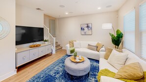 Ein modernes Wohnzimmer mit Flachbildfernseher, einem grauen Pouf, einem weißen Sofa und einem blauen Teppich mit Muster.