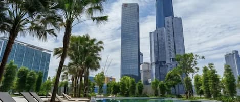 A poolside area with lounge chairs, palm trees, and a city skyline in the background.