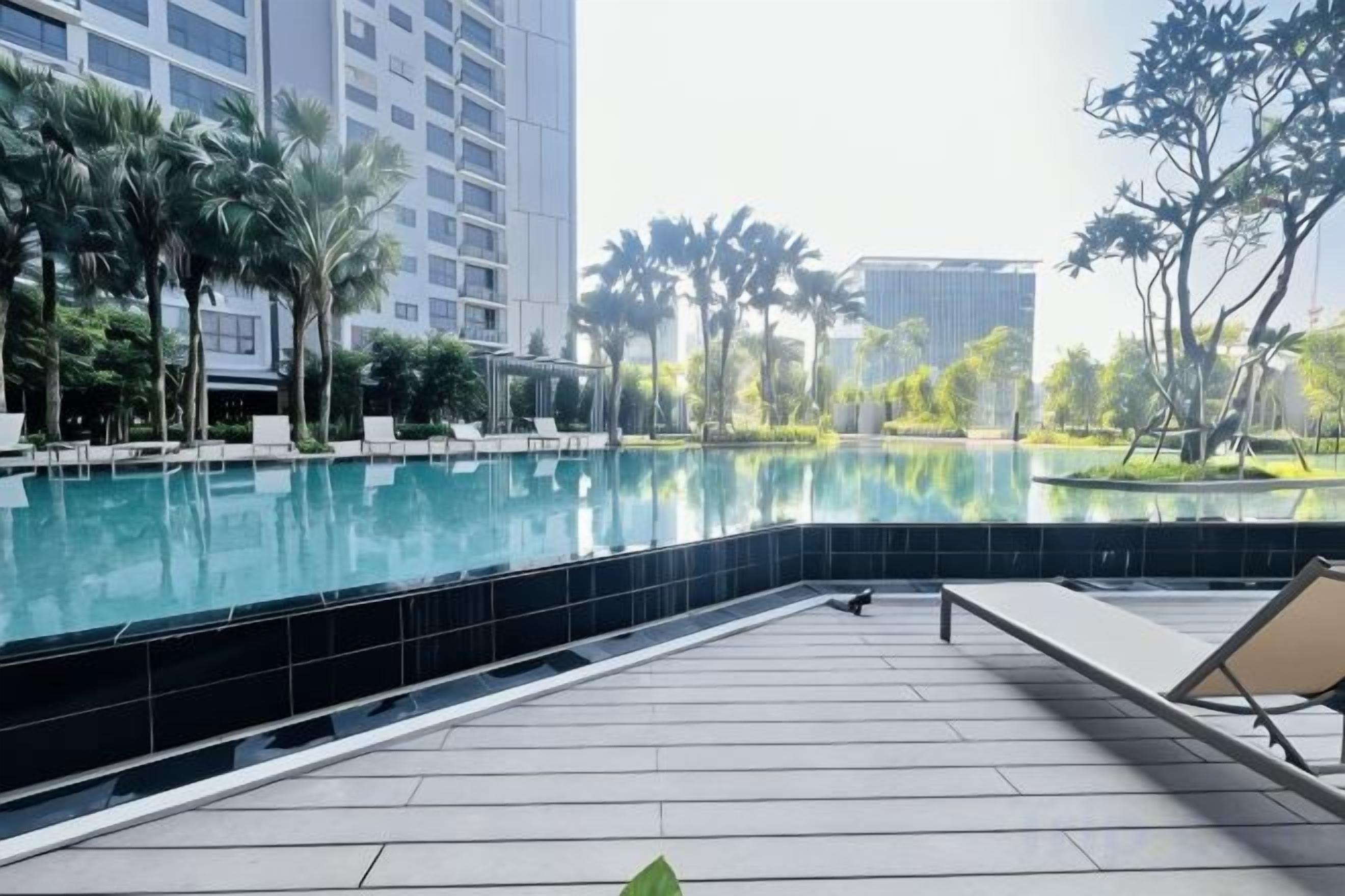 A swimming pool with lounge chairs and a tiled deck, surrounded by palm trees and modern buildings.