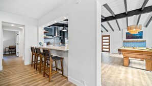 Una cuina moderna amb armaris blancs, una zona de bar amb tamborets de fusta i billar en una habitació espaiosa amb sòl de fusta.