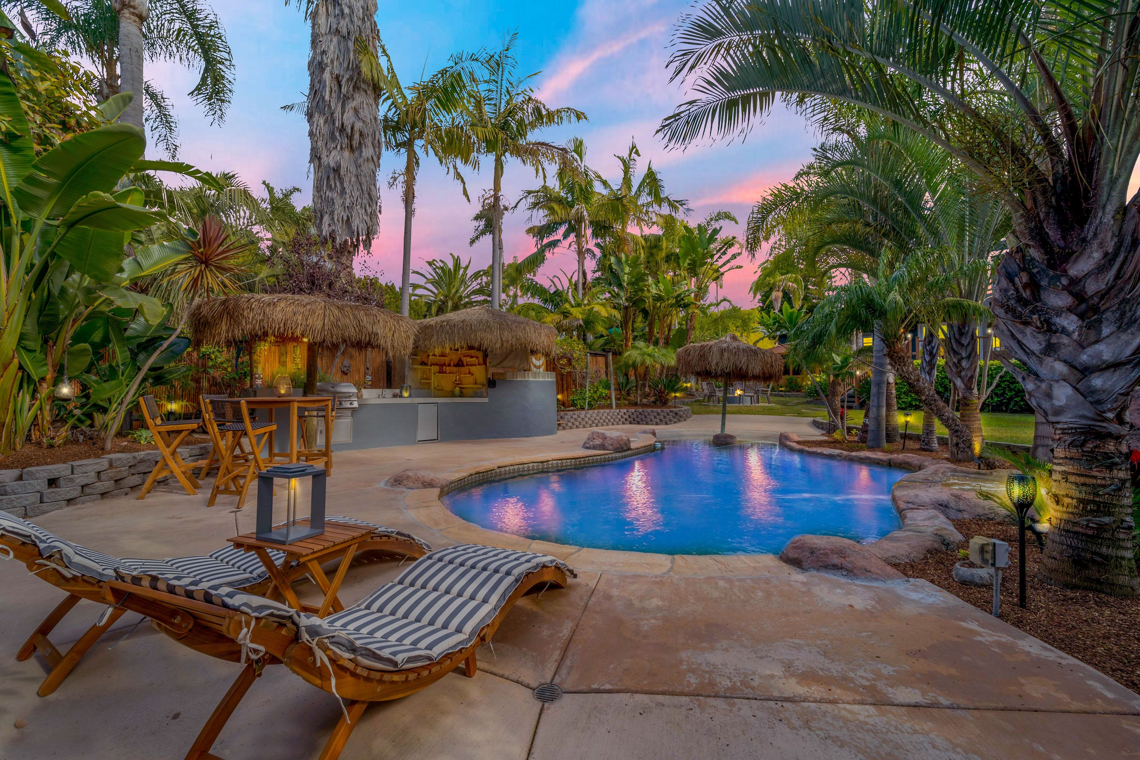 A pool area with lounge chairs, a barbecue, and thatched-roof structures.