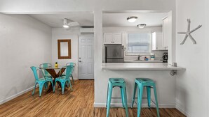 A modern kitchen with white cabinets, a stainless steel refrigerator, and a window with blinds. There is a dining area with a wooden table and turquoise chairs.
