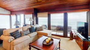 A living room with a wooden ceiling, a sofa with patterned cushions, a coffee table with candles, and a TV on a wooden cabinet. - Luxury Beachfront Getaway - Main House (Oceanside)
