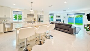 A modern kitchen with white cabinetry, a central island with bar stools, and a dining area with a gray sofa. - Enchanting Cottage by the Sea (Carlsbad)
