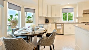 A modern kitchen with white cabinetry, a dining area with a round table and chairs, and large windows with views of palm trees. - Enchanting Cottage by the Sea (Carlsbad)
