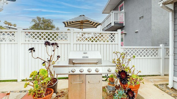 A barbecue grill in a backyard with a white fence, potted plants, and a house in the background. - The Strand Cottage Beachfront (Oceanside)