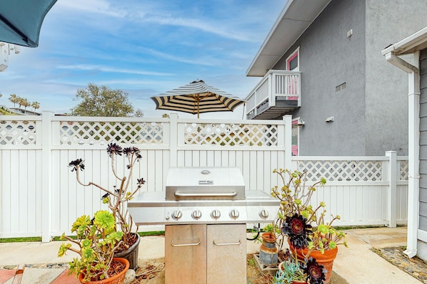 A barbecue grill in a backyard with a white fence, potted plants, and a house in the background.