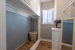 A laundry room with a wooden floor, a laundry basket, a white cabinet, and a window with blinds.