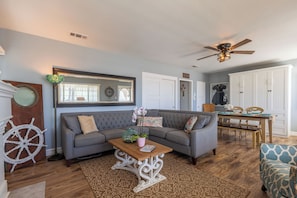 A living room with a sectional sofa, a wooden coffee table, a decorative mirror, a ceiling fan, and a dining area with a table and chairs.