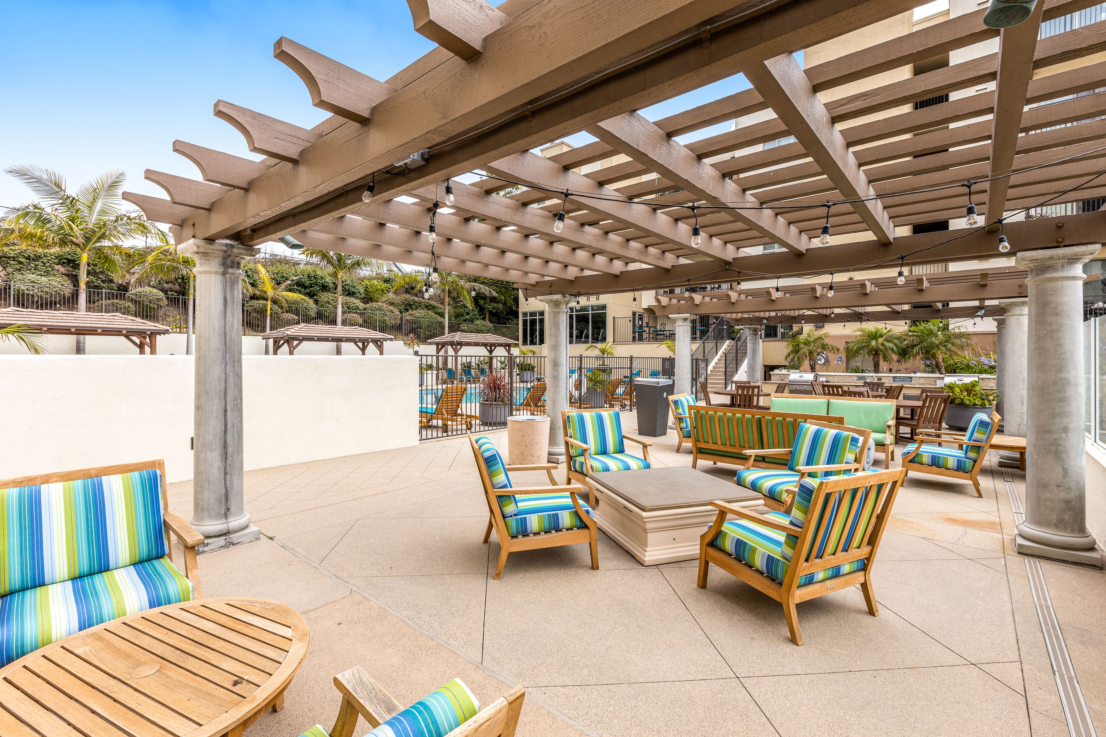 An outdoor patio area with wooden furniture, a pergola, and a pool in the background.