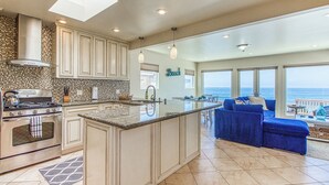 A spacious kitchen with a large island, a dining area with a view, and a living room with a blue sofa. - Ocean Beach Cliffs Sanctuary Oceanfront (San Diego)