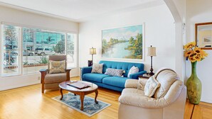 A living room with a blue sofa, beige armchair, and a white sofa. There is a painting of a river landscape on the wall, a lamp, and a vase with sunflowers.