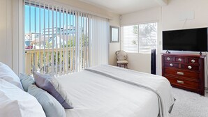 A bedroom with a large bed, a dresser, a chair, a TV, and a balcony with blinds.