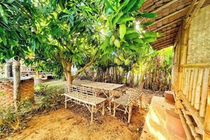 Un'area salotto esterna con tavolo e sedie sotto un albero, immersa in un edificio rustico con tetto di paglia.