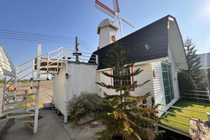 A small building with a white exterior, a black roof, and a red and white windmill in the background. - khao Thung Benja HomeStay (Phayuha Khiri)