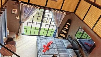 A room with a bed, a chair, a small table, and a view of a green outdoor area through large windows.