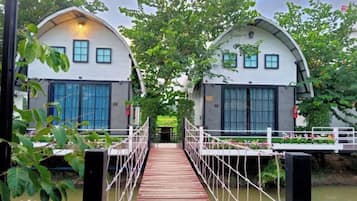 A wooden bridge leading to a row of white buildings with large windows, surrounded by greenery.
