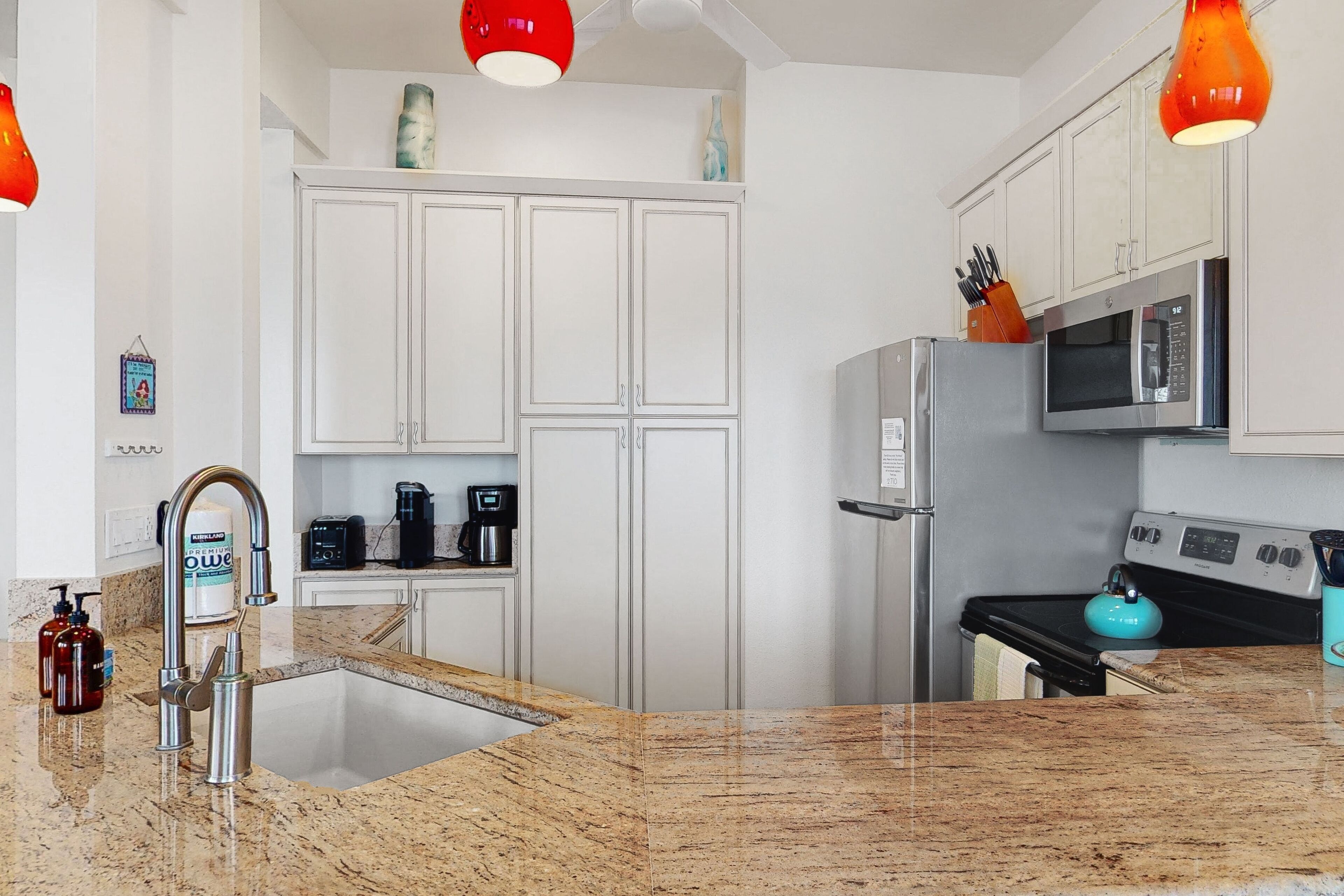 Una cocina moderna con armarios blancos, electrodomésticos de acero inoxidable, encimera de granito y lámparas colgantes rojas.