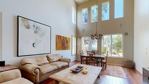 Amplia sala de estar con un sofá color beige, una mesa de centro y un comedor con sillas y una mesa de madera. Amplias ventanas dejan entrar luz natural y ofrecen vistas al exterior.