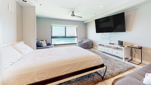 A modern bedroom with a large bed, a TV mounted on the wall, a ceiling fan, and a view of the ocean through a window. - Blue Wave Penthouse Oceanview (San Diego)
