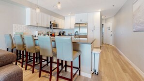 A modern kitchen with white cabinets, a central island with bar stools, stainless steel appliances, and pendant lighting. - Blue Wave Penthouse Oceanview (San Diego)