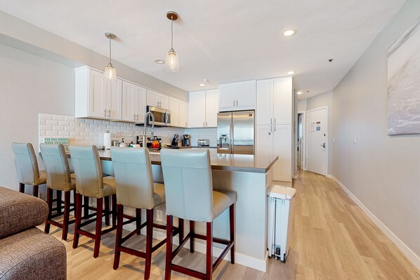 A modern kitchen with white cabinets, a central island with bar stools, stainless steel appliances, and pendant lighting. - Blue Wave Penthouse Oceanview (San Diego)