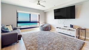 A modern living room with a large window, a grey sofa, a bean bag, a flat-screen TV, and a ceiling fan. - Blue Wave Penthouse Oceanview (San Diego)