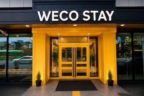 L’entrée d’un hôtel appelé WECO STAY, avec une porte en verre et un paillasson noir devant.