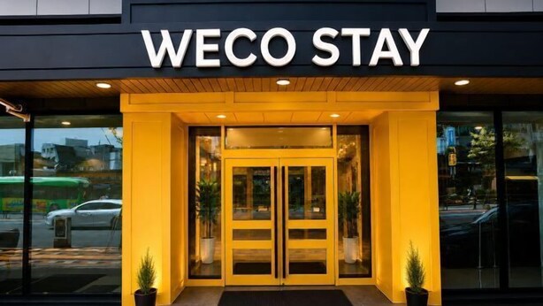 Der Eingang eines Hotels mit einem 'WECO STAY' Schild, Glastüren und Pflanzkübeln.
