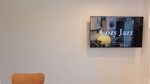 Ein Zimmer mit an der Wand montiertem Fernseher, der 'Cozy Jazz' zeigt, einem weißen Bett und einem braunen Sessel.