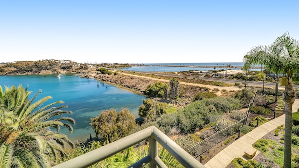 Paisaje costero con un lago azul cristalino, un puente a lo lejos y un camino que conduce a un edificio con barandilla.