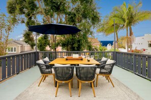 Een terras buiten met een houten tafel en stoelen, een blauwe parasol en uitzicht op de oceaan.
