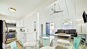 Una cocina moderna con gabinetes blancos, un microondas y un horno. Hay un comedor con una mesa de vidrio y sillas, y una sala de estar con un sofá y una televisión.
