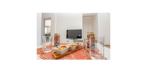 Un salon moderne avec un téléviseur à écran plat, une table basse en verre sur laquelle sont disposés des en-cas, et un chemin de table rouge à motifs.