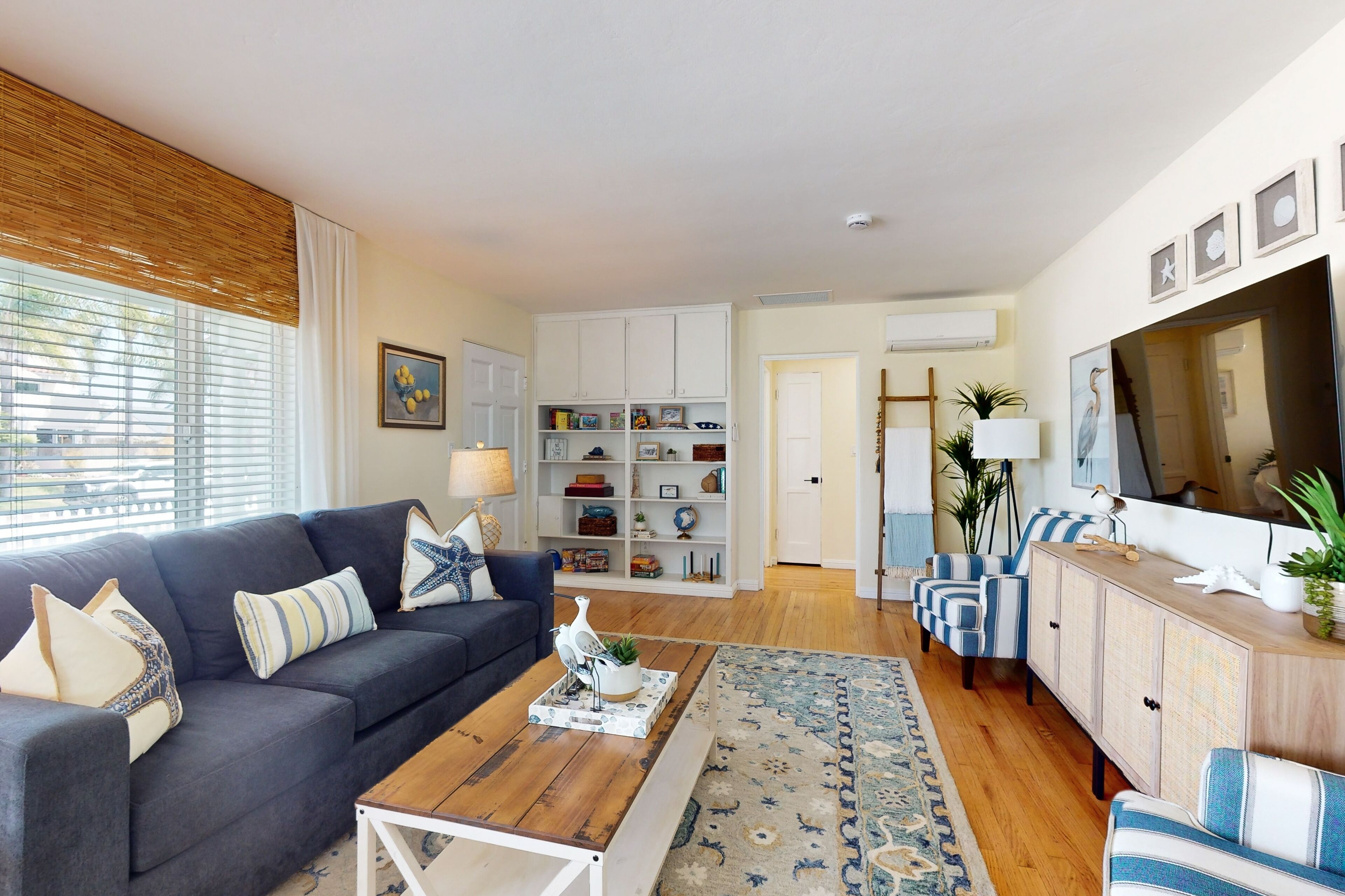 A living room with a dark grey sofa, a wooden coffee table, and a patterned rug. There is a white cabinet with shelves, a television, and a potted plant.