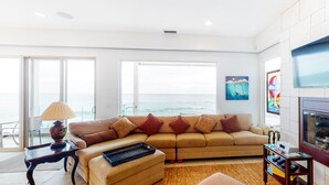 A spacious living room with a large window overlooking the ocean, a comfortable sofa, a small table with a lamp, and a fireplace. - Luxury Oceanfront Home - Lower (Oceanside)