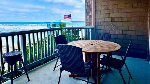 A balcony with a round wooden table and four chairs, overlooking the ocean with a view of the beach and a flag.