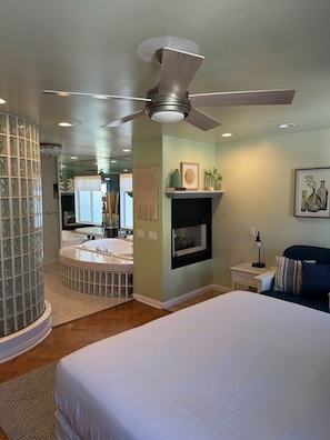 A bedroom with a bed, a ceiling fan, a fireplace, and a bathroom with a glass shower enclosure.