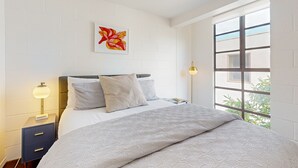 Ein Schlafzimmer mit Bett, Nachttisch, Lampe und einem Fenster mit Blick auf Grünflächen.